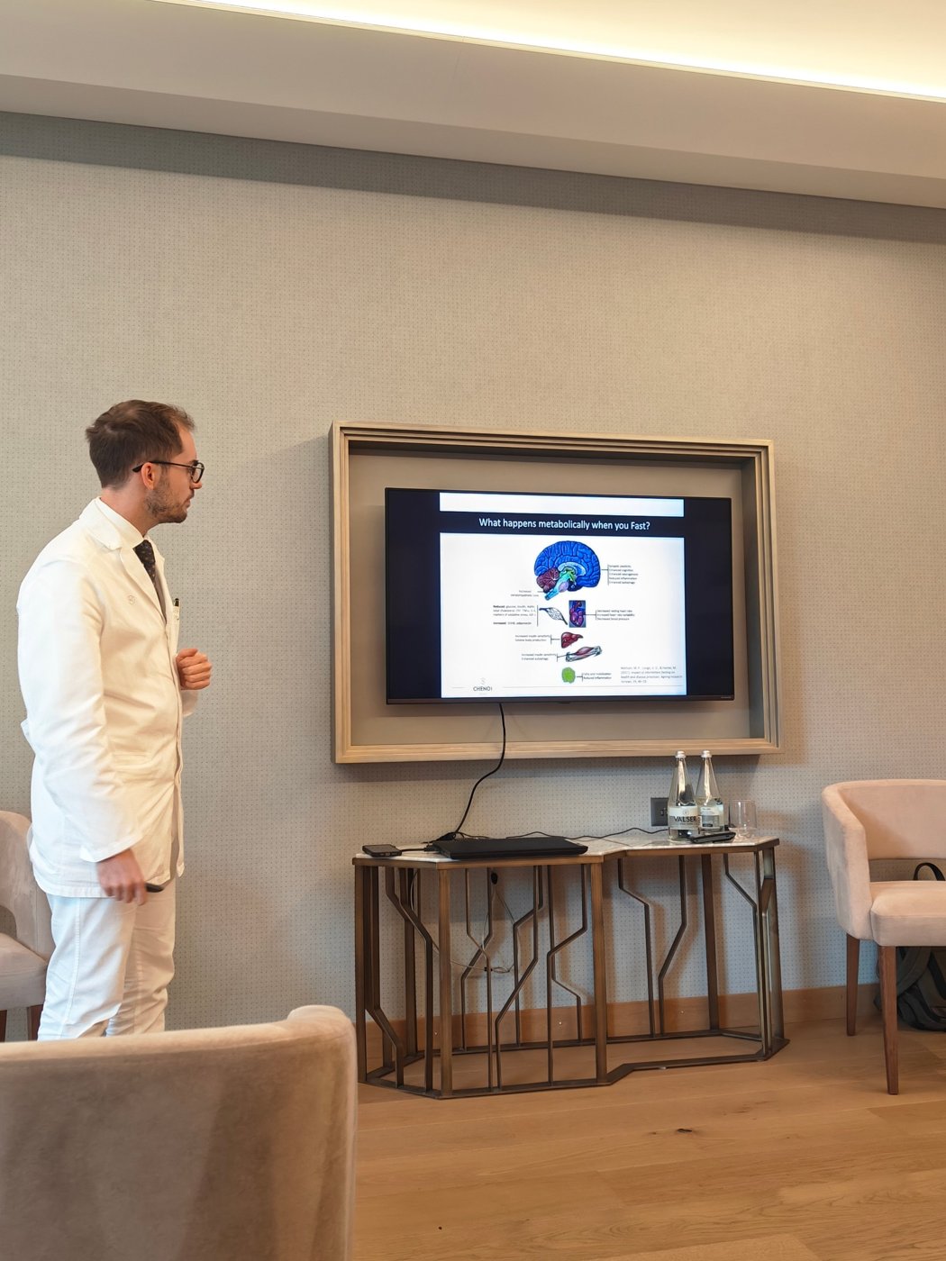A Chenot doctor in a white coat giving a talk to guests in front of a slide titled What happens metabolically when you Fast?