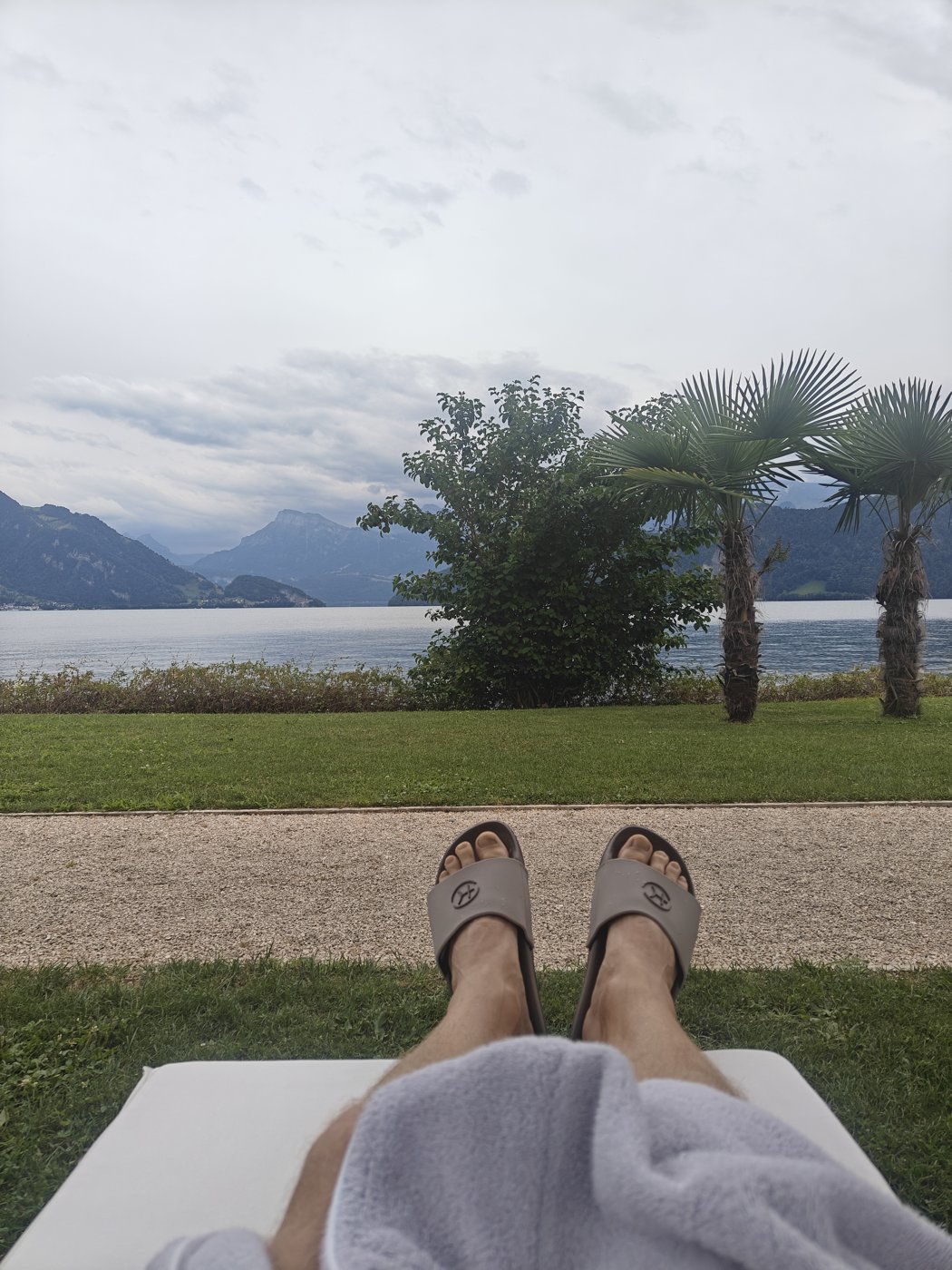 Feet up on a lounger on the lawn at Chenot Palace Weggis, looking over Lake Lucerne with palms and mountains