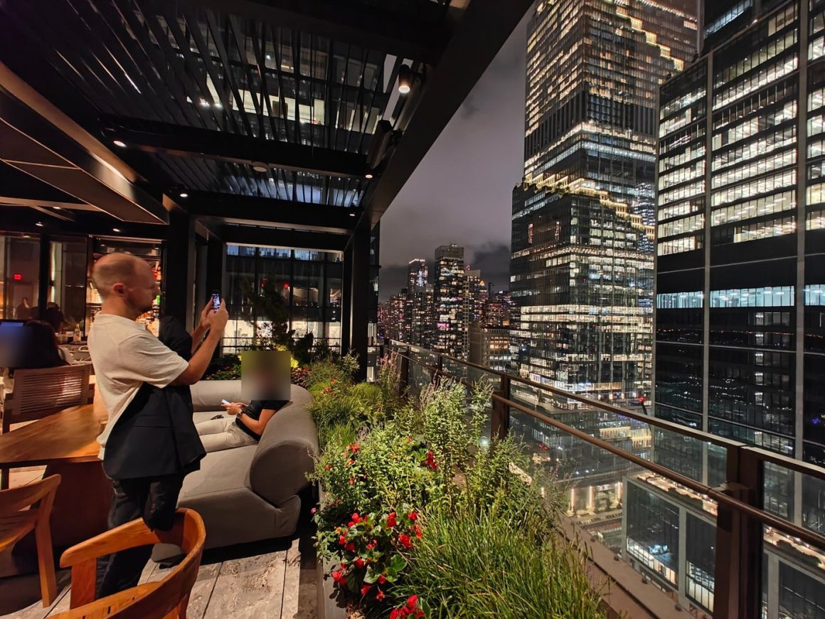The Electric Lemon terrace at night, with planters along a glass railing and the lit towers of Hudson Yards, a guest photographing the view