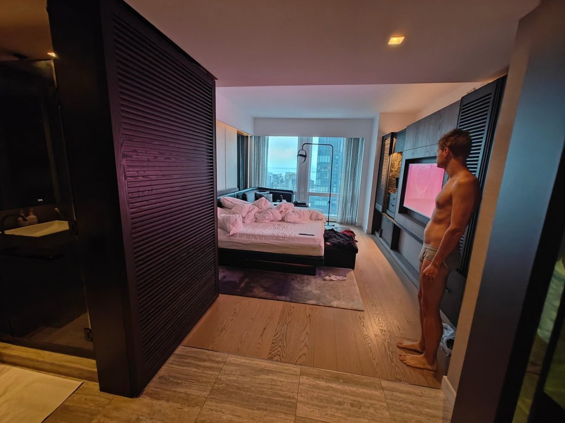 High-floor Equinox guest room with dark slatted wood partitions, an unmade bed and a floor-to-ceiling window over Manhattan towards the Hudson, a guest standing by the TV wall